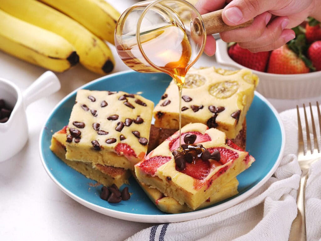 A hand pours syrup over slices of sheet pan pancakes on a blue plate, with bananas and strawberries in the background.