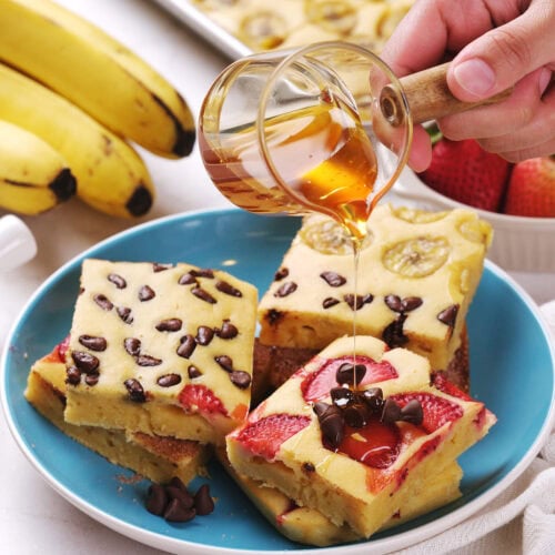 A hand pours syrup over stacked pieces of sheet pan pancakes on a blue plate.