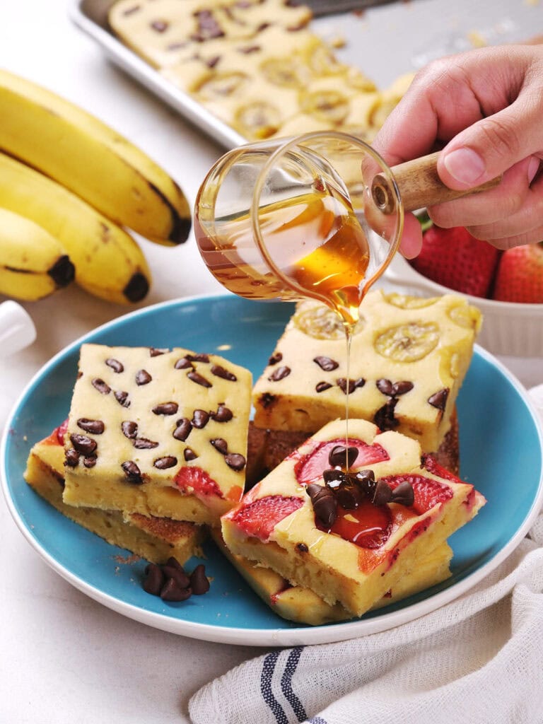 A hand pours syrup over stacked pieces of sheet pan pancakes on a blue plate.