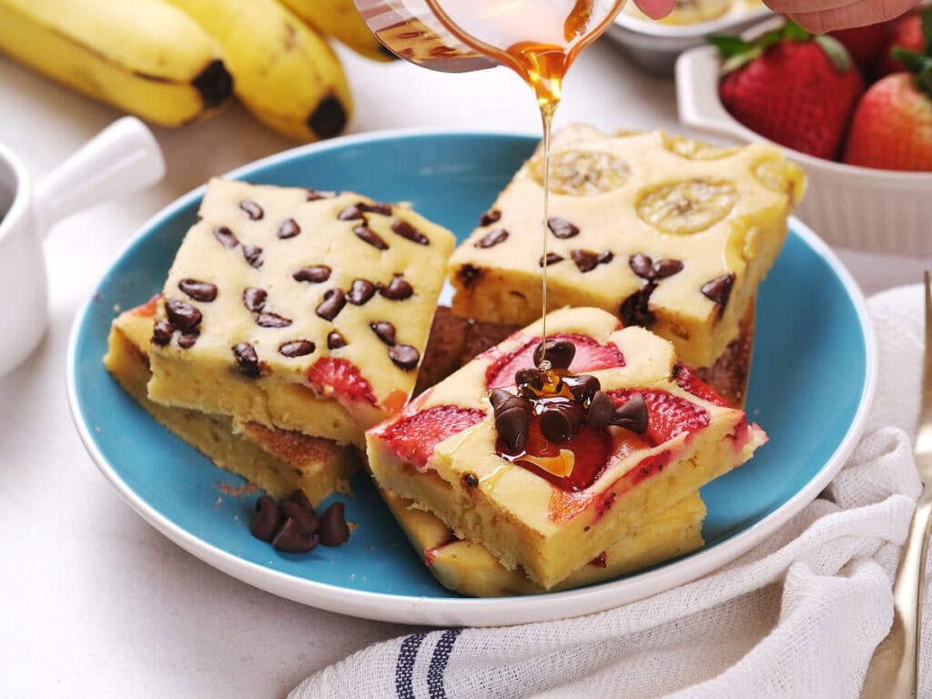 A hand pours syrup onto stacked sheet pan pancakes.