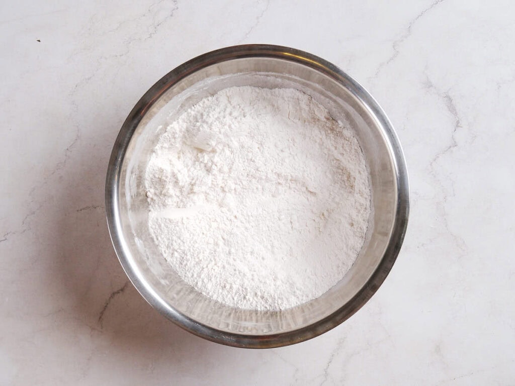 A stainless steel bowl filled with white flour sits on a light-colored marble surface.