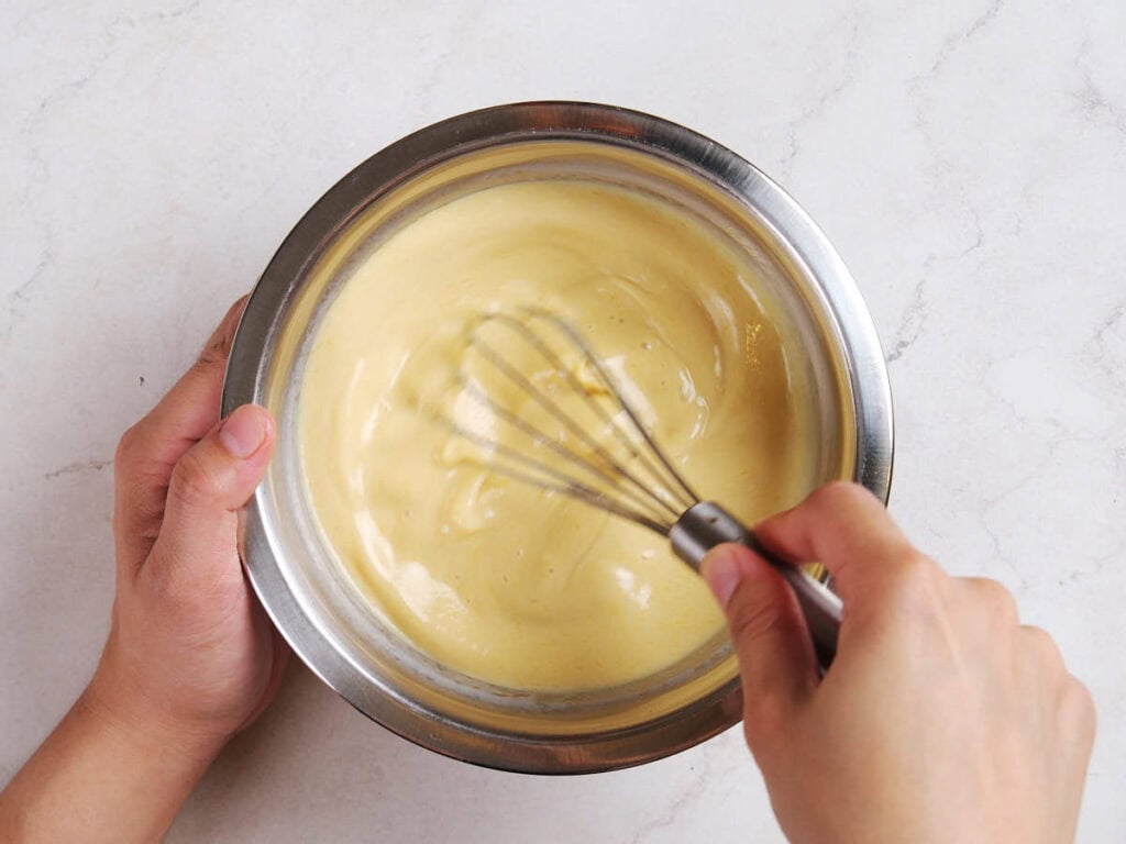 A person whisking creamy yellow batter in a metal bowl on a light-colored surface.