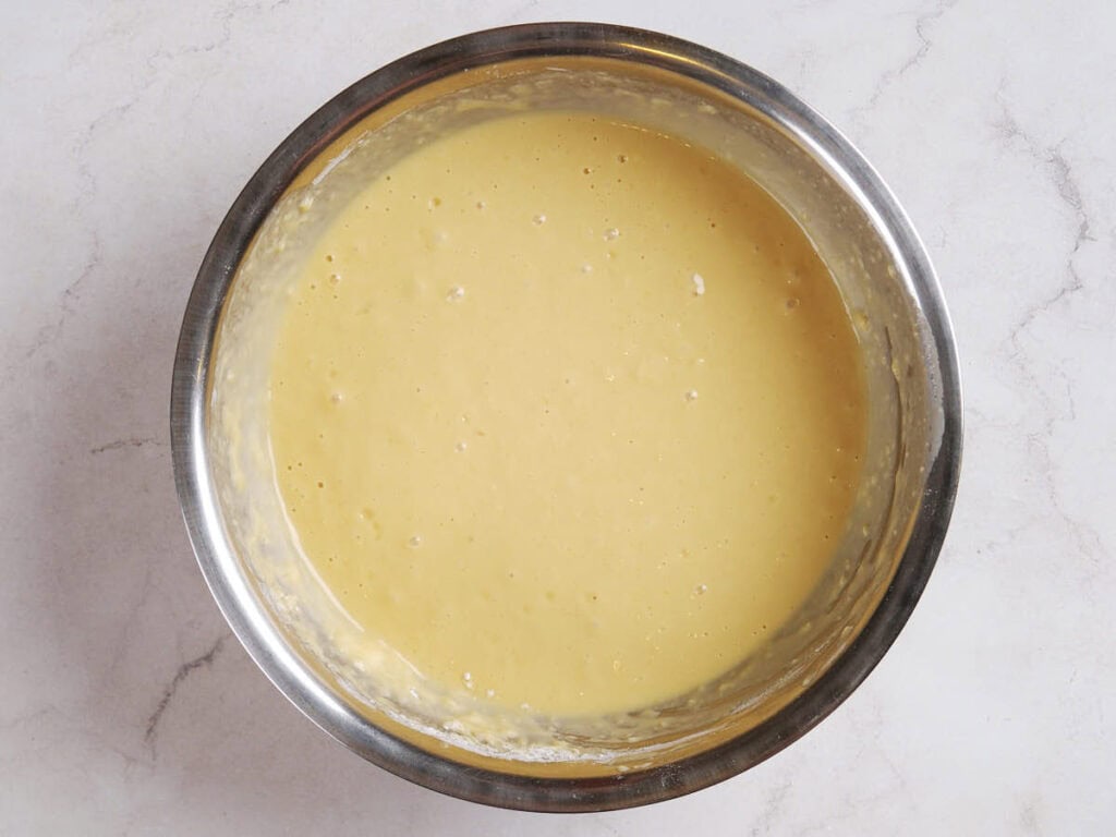 A stainless steel bowl filled with smooth, light yellow batter sits on a white marble surface.