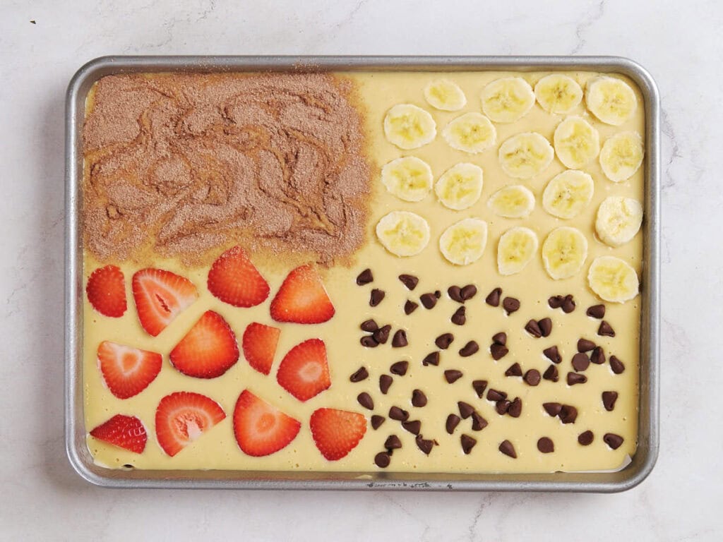 A rectangular pan of unbaked batter topped with four sections: cinnamon sugar, banana slices, chocolate chips, and sliced strawberries, on a light surface.