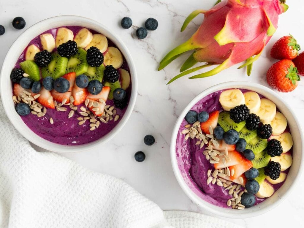 Two smoothie bowls topped with sliced bananas, kiwi, strawberries, blackberries, blueberries, and sunflower seeds.