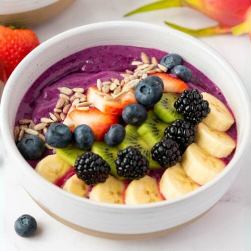 A smoothie bowl topped with sliced banana, kiwi, strawberries, blackberries, blueberries, and sunflower seeds.