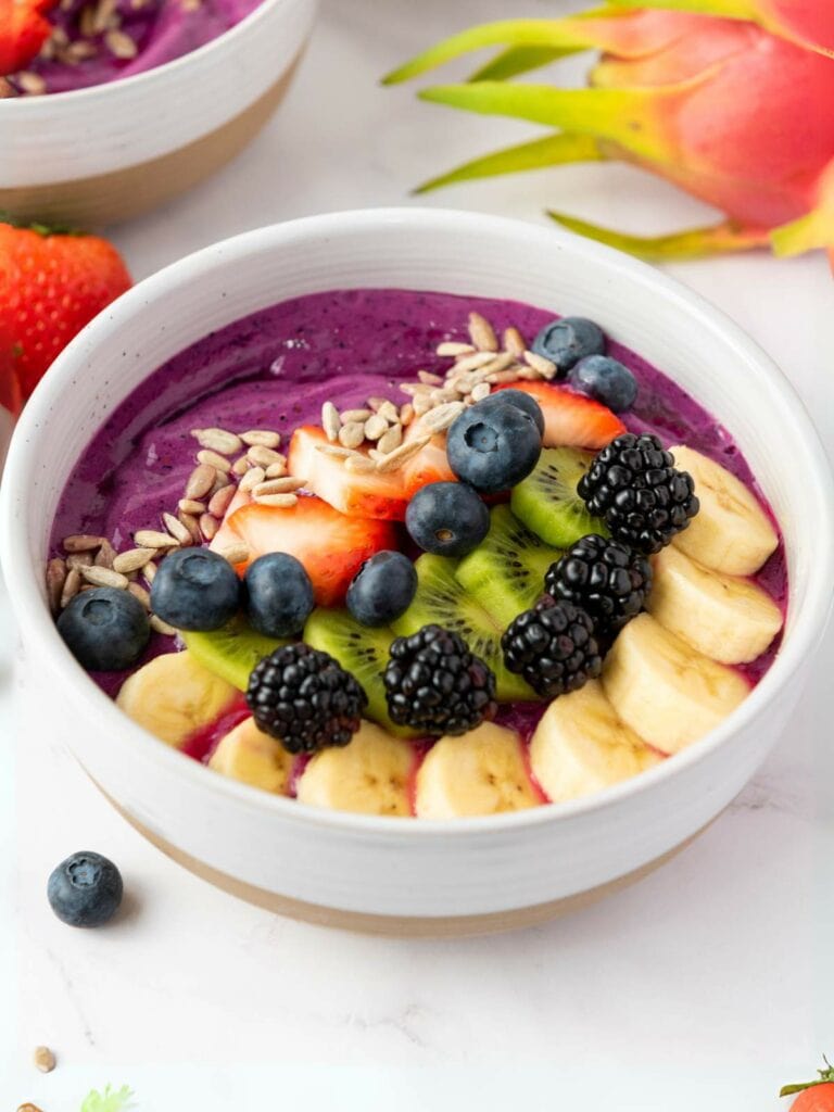A smoothie bowl topped with sliced banana, kiwi, strawberries, blackberries, blueberries, and sunflower seeds.