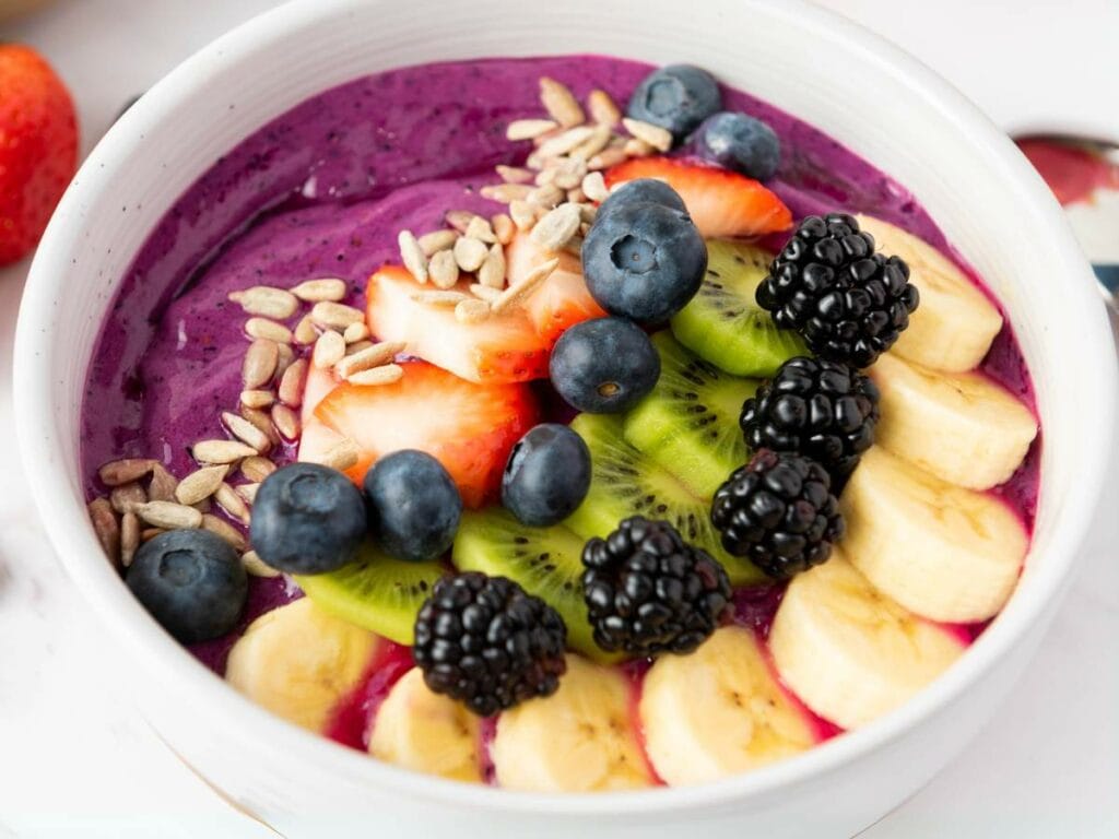 A smoothie bowl topped with sliced banana, kiwi, strawberries, blackberries, blueberries, and sunflower seeds in a white bowl.