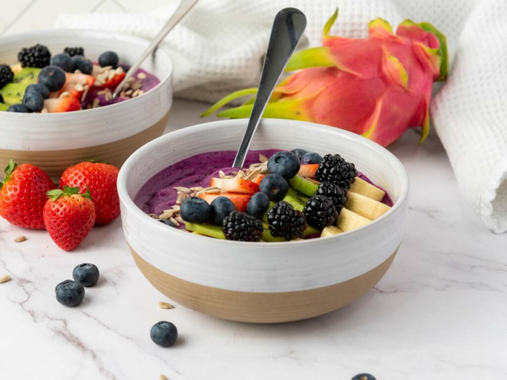 Two bowls of smoothie topped with banana, kiwi, blackberries, blueberries, and strawberries on a marble surface.