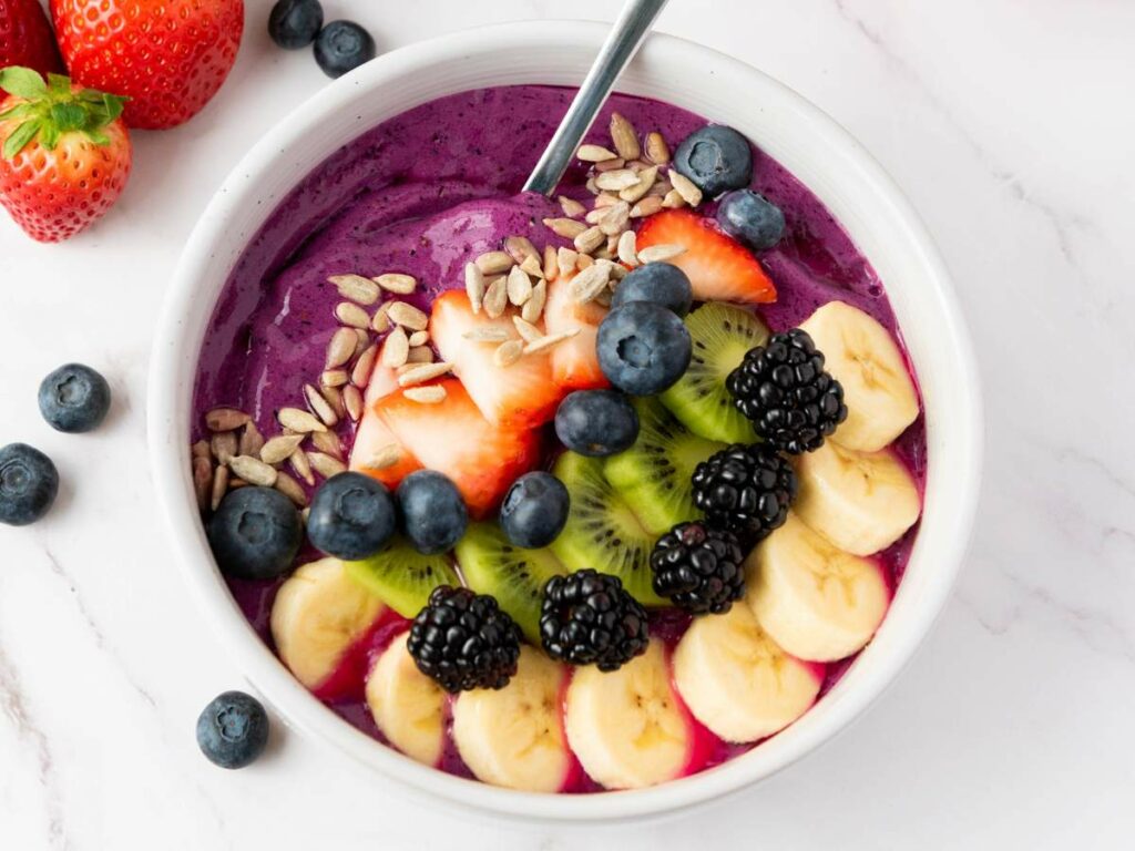 A smoothie bowl topped with sliced banana, kiwi, strawberries, blackberries, blueberries, and sunflower seeds.