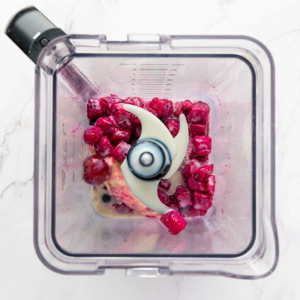 Top view of a blender containing frozen red berries, banana, and a small amount of liquid, ready to be blended.