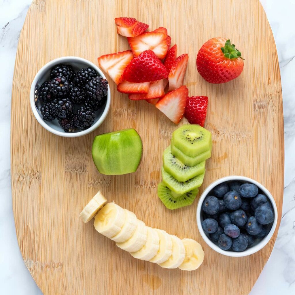 A wooden cutting board with bowls of blackberries and blueberries, sliced strawberries, a whole strawberry, chopped kiwi, green apple chunk, and sliced banana.