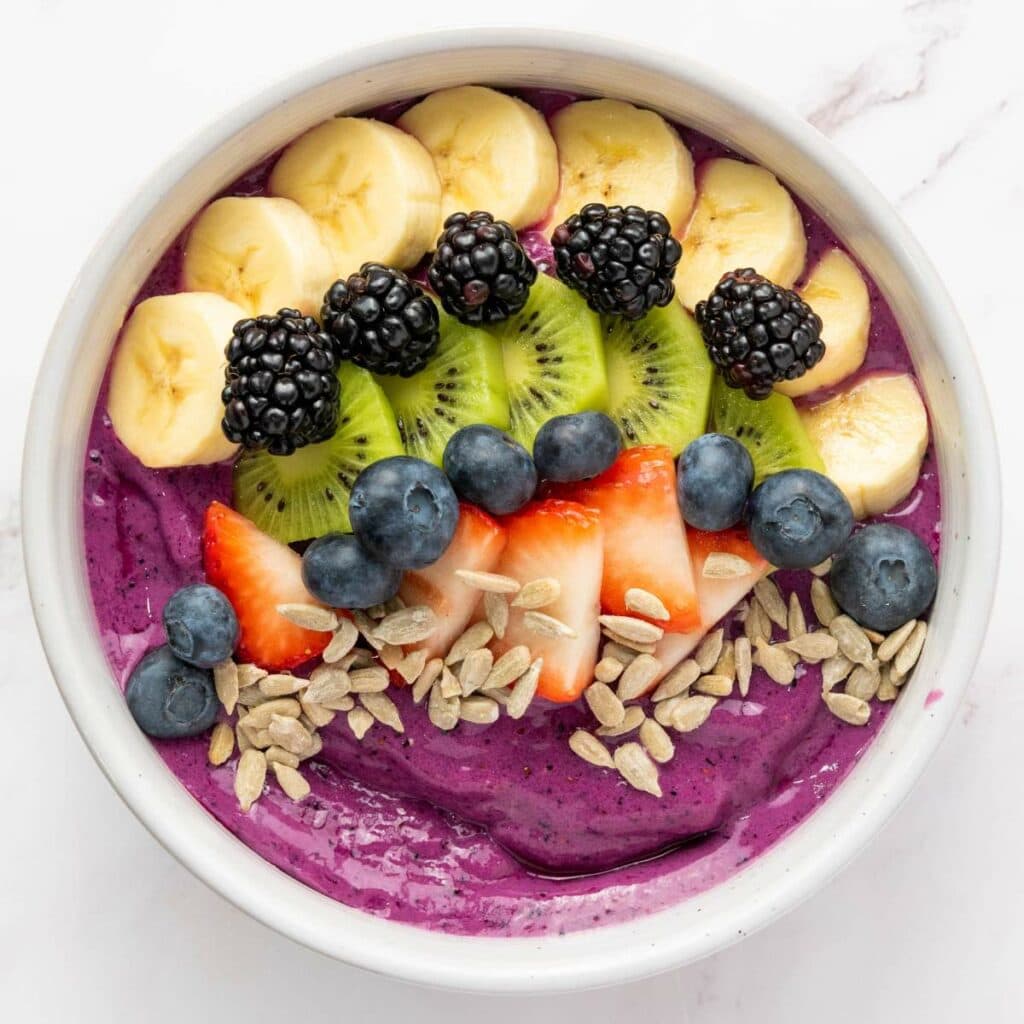 A bowl of purple smoothie topped with sliced bananas, kiwi, blackberries, blueberries, strawberries, and sunflower seeds.