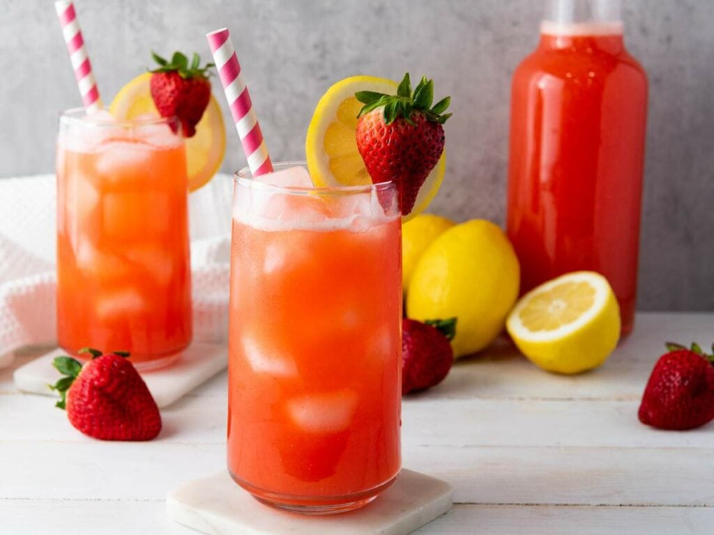 Two glasses of strawberry lemonade with ice, garnished with sliced lemon and whole strawberries.