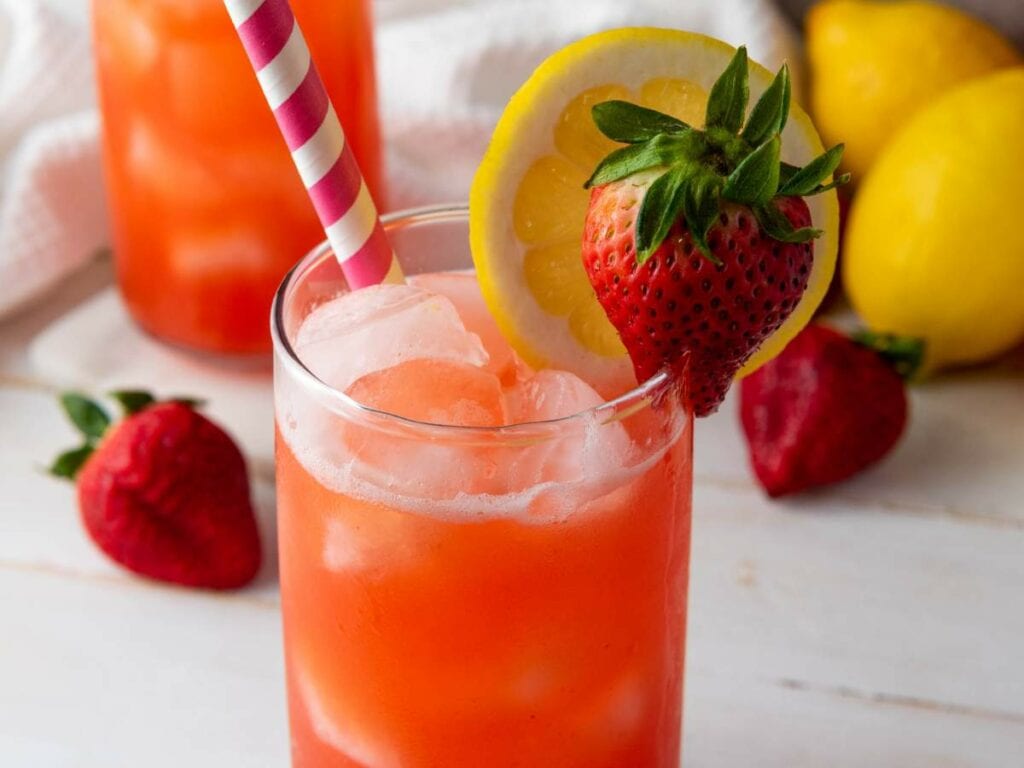 A glass of red drink with ice, garnished with a strawberry and lemon slice, and a striped straw. Lemons and strawberries are in the background.