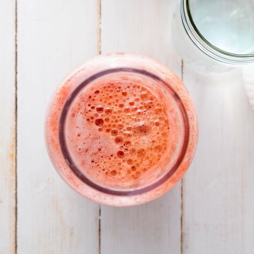 Top view of a glass jar filled with frothy pink strawberry-infused liquid on a light wooden surface. A partially visible empty jar is beside it.