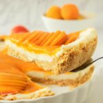 A slice of apricot tart with a crumb crust and sliced fresh apricot topping is being lifted from a white pie dish.