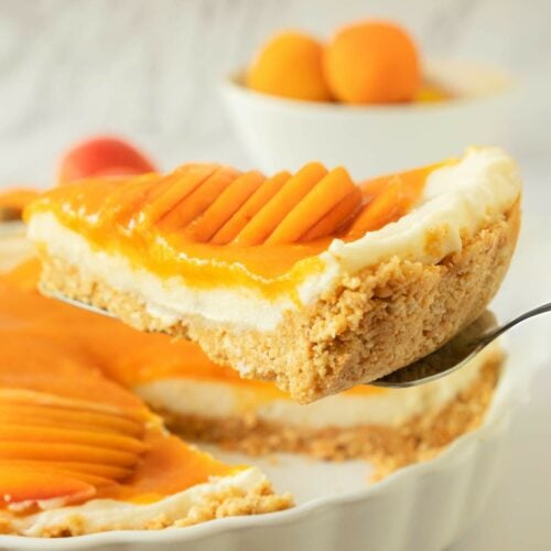 A slice of apricot tart with a crumb crust and sliced fresh apricot topping is being lifted from a white pie dish.
