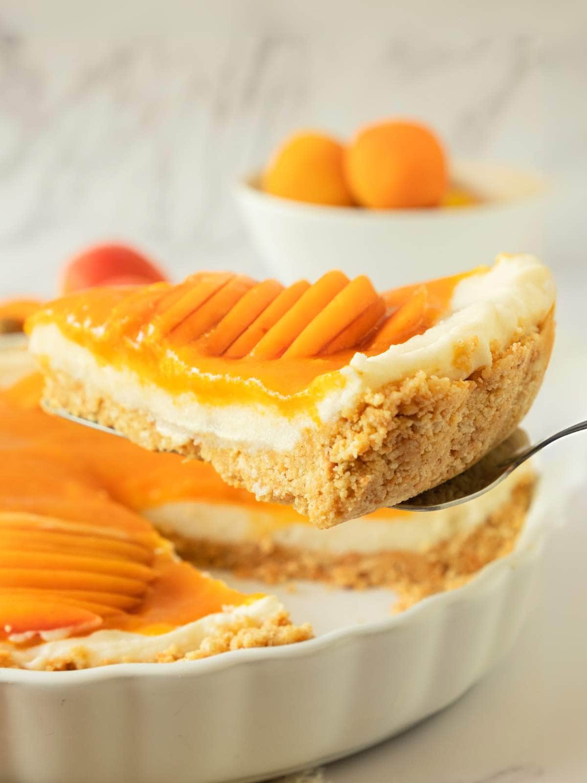 A slice of apricot tart with a crumb crust and sliced fresh apricot topping is being lifted from a white pie dish.