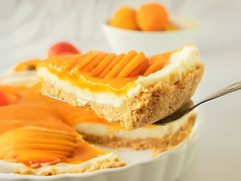 A slice of apricot tart with a crumbly crust is being lifted from a pie dish.