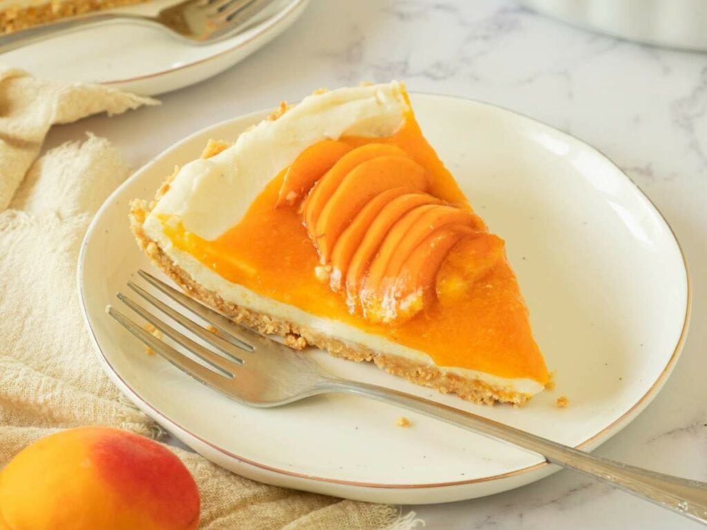 A slice of apricot tart garnished with fresh apricot slices sits on a white plate with a fork beside it.