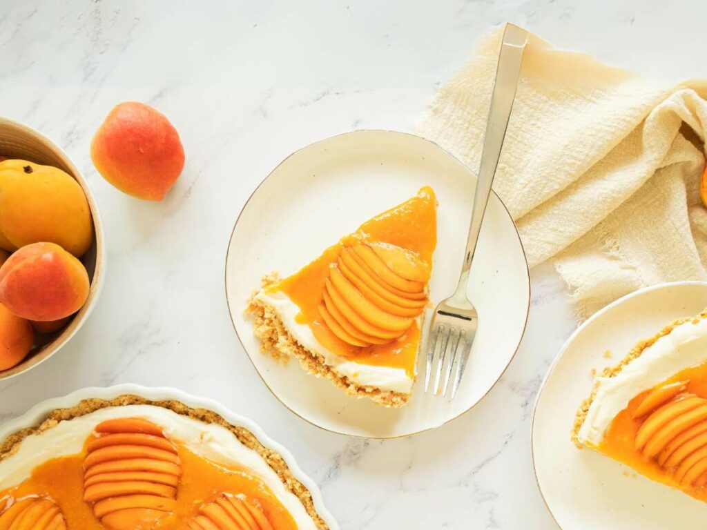 A slice of apricot tart with a fork on a plate, next to a whole tart, fresh apricots, and a beige napkin on a marble surface.