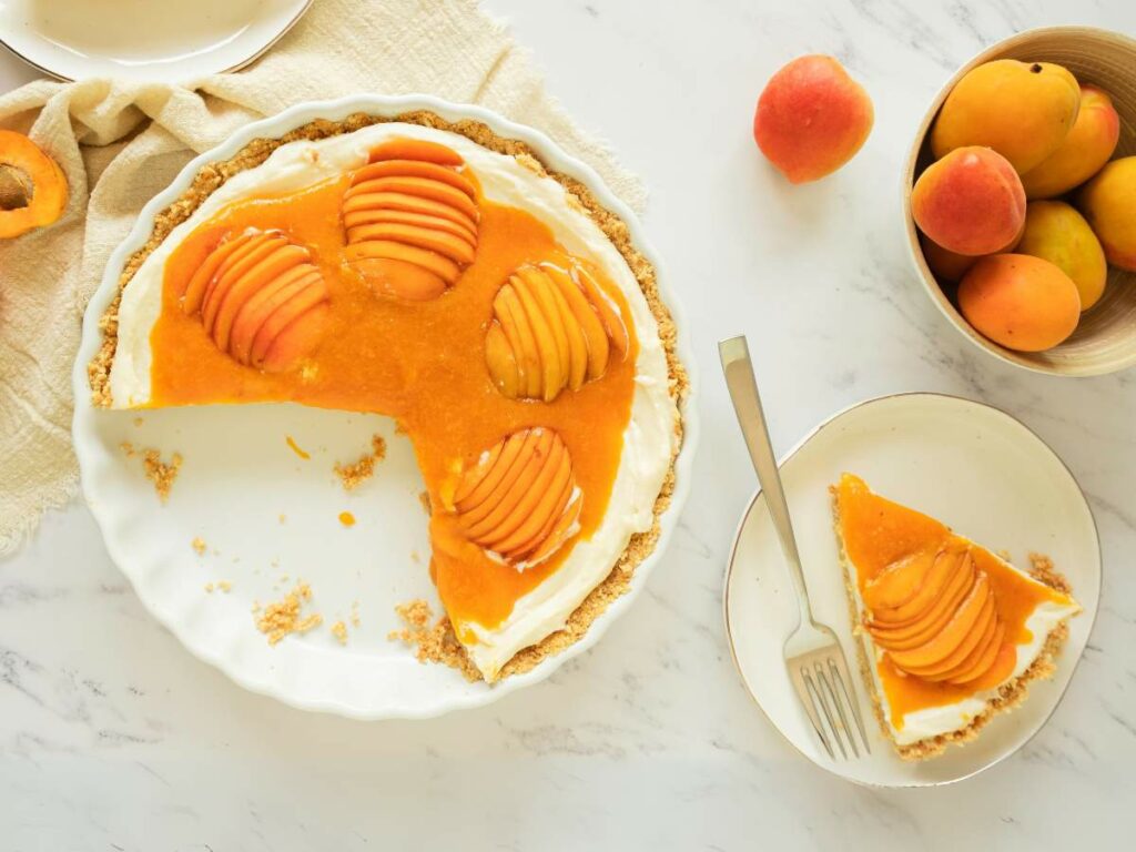 A partially sliced apricot tart with a creamy filling on a white plate, next to a single served slice.