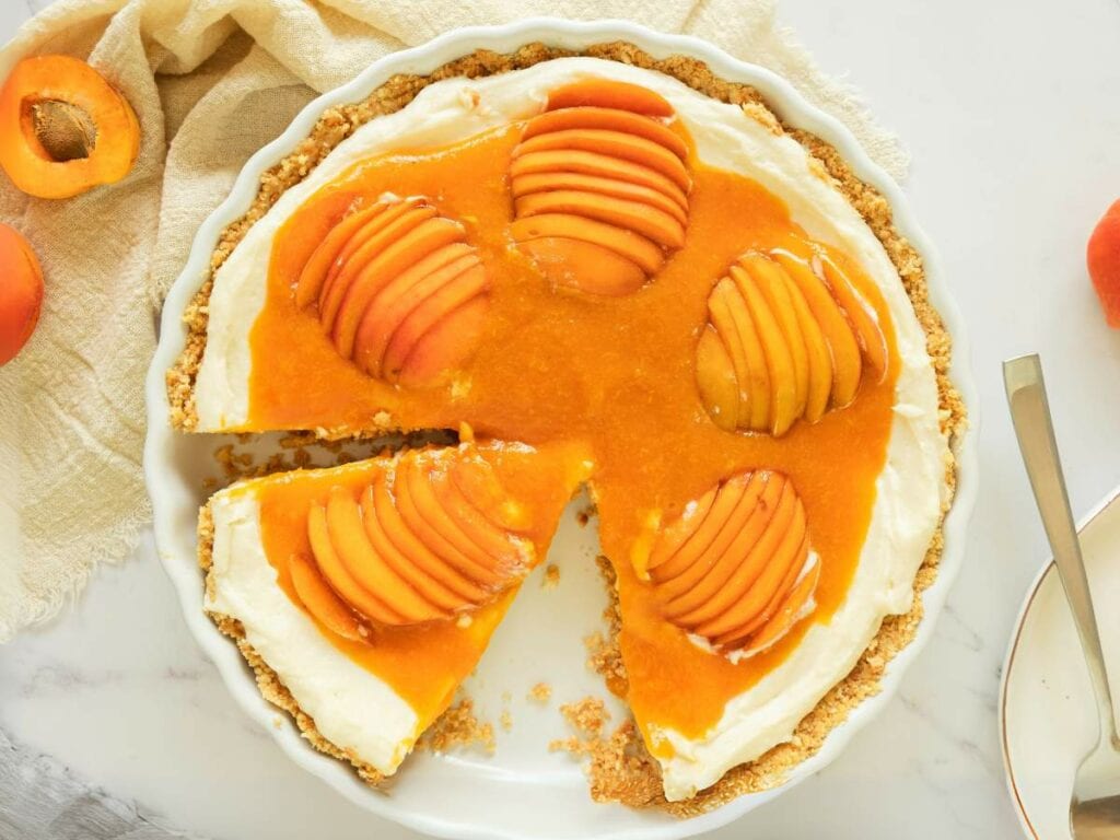 A sliced apricot tart with a cream filling and a crumb crust, topped with fanned fresh apricots and a glossy apricot glaze, on a white plate.
