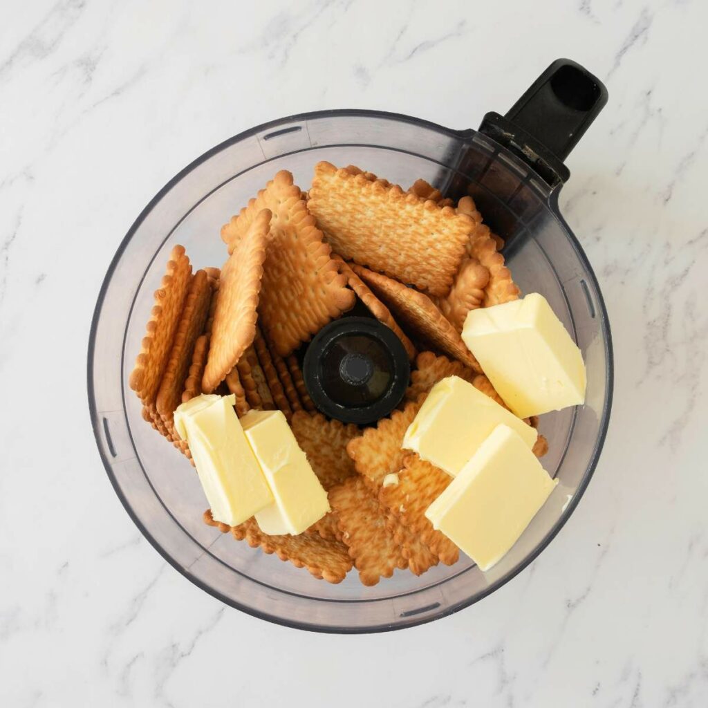 Rectangular biscuits and three chunks of butter in a food processor bowl on a white marble surface.