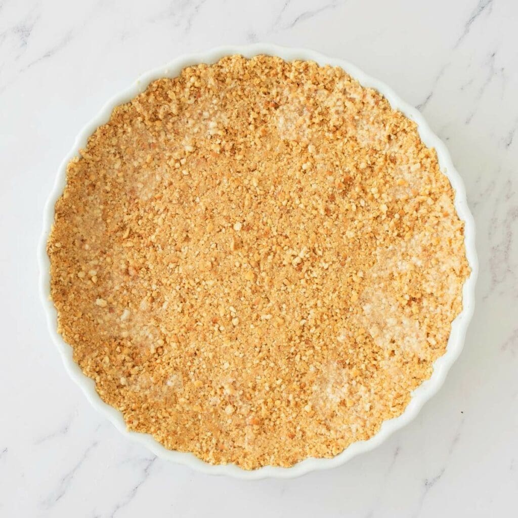 A round pie dish filled with an even layer of crushed biscuit or graham cracker crust on a white marble surface.