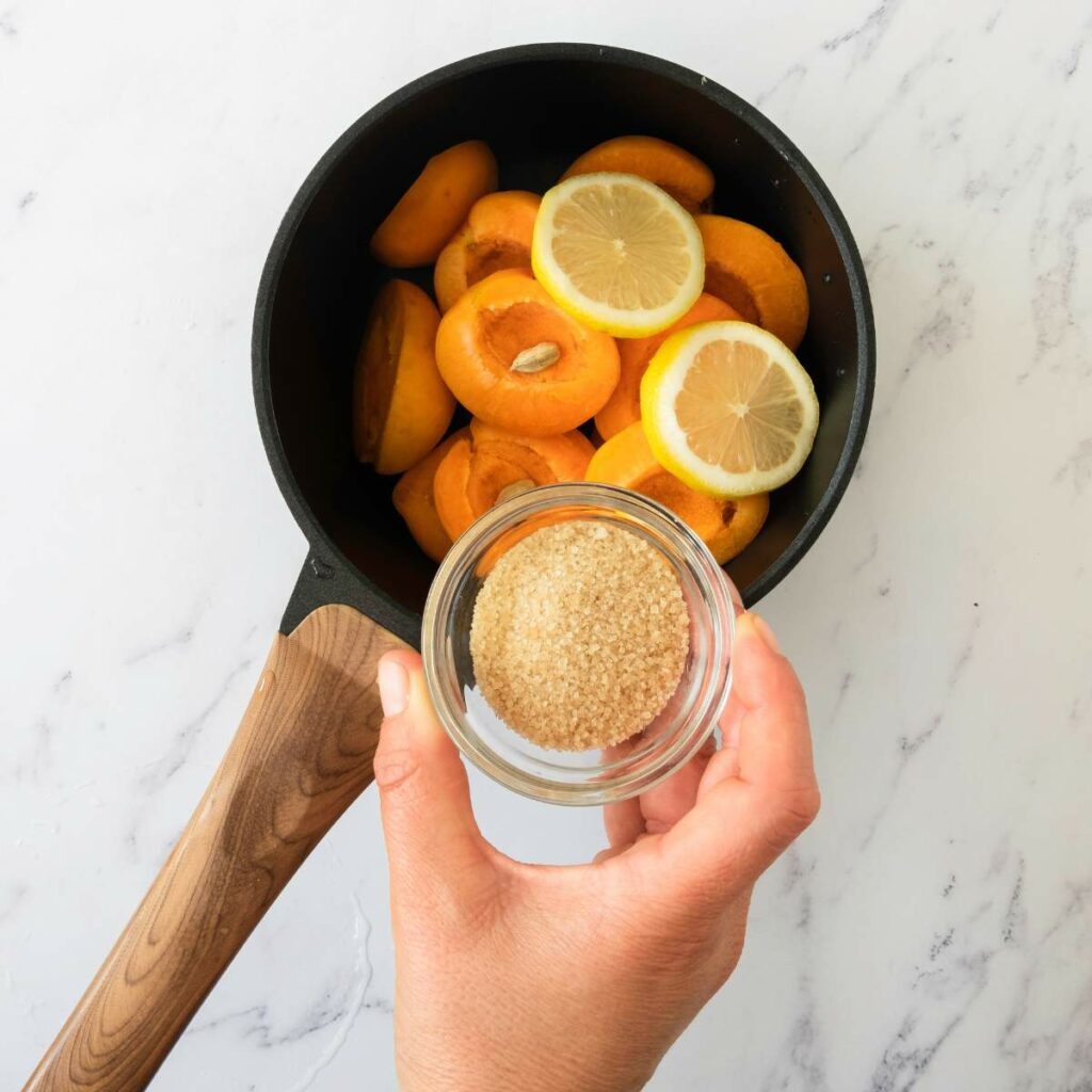 A hand holds a small bowl of brown sugar over a saucepan filled with halved apricots and lemon slices on a white marble surface.