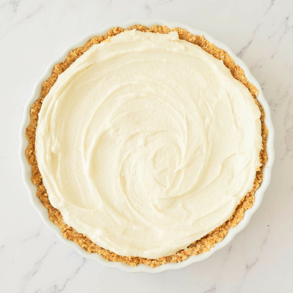 A pie dish filled with a graham cracker crust and a smooth, creamy white filling on a white marble surface.