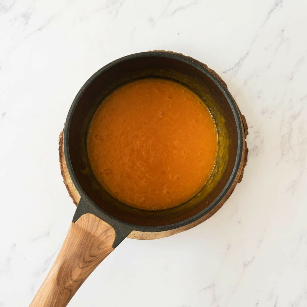 A small saucepan with a wooden handle containing orange-colored sauce, placed on a round wooden trivet on a white marble surface.