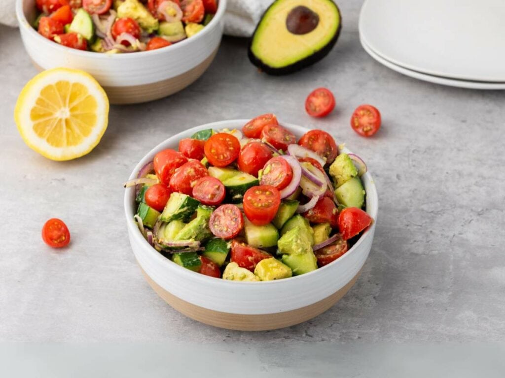 A bowl of salad with cherry tomatoes, avocado, red onion, and cucumber sits on a grey surface next to a lemon slice and a halved avocado.