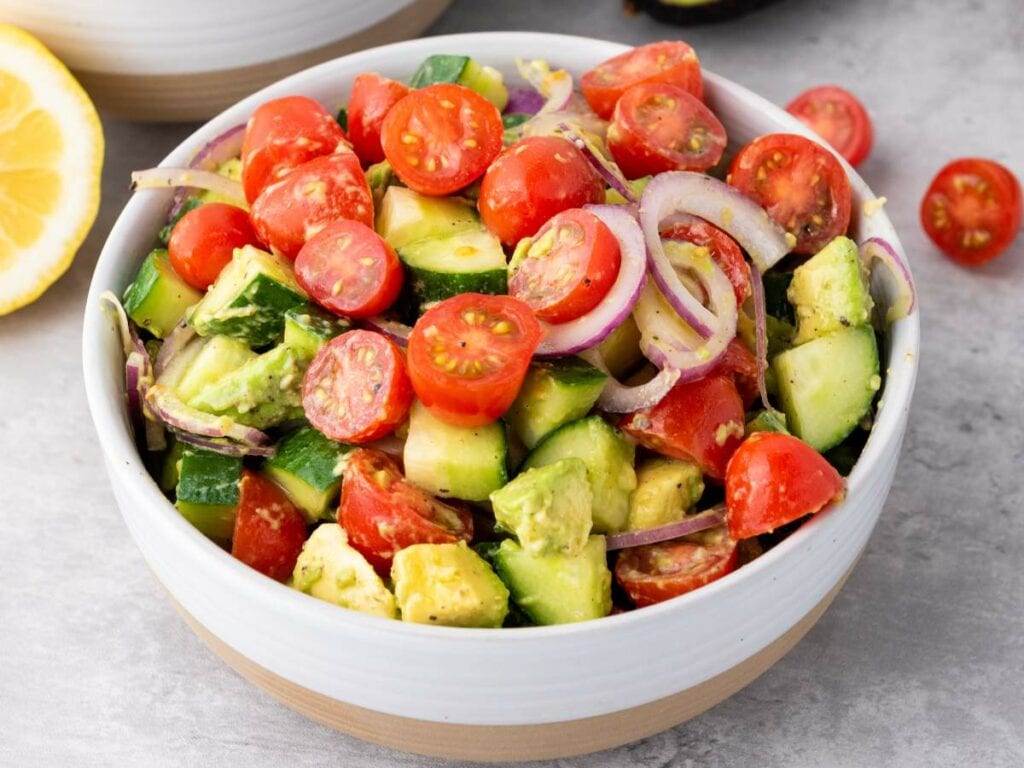 A white bowl filled with a salad of cherry tomatoes, cucumber, avocado, and red onion slices on a gray surface.