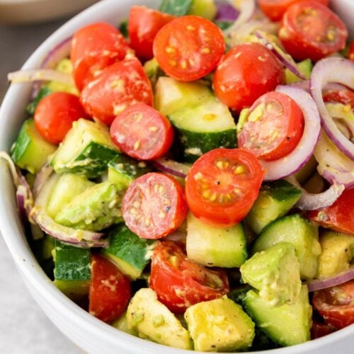 A bowl of salad containing sliced cherry tomatoes, cucumber, avocado, and red onion with a light dressing.