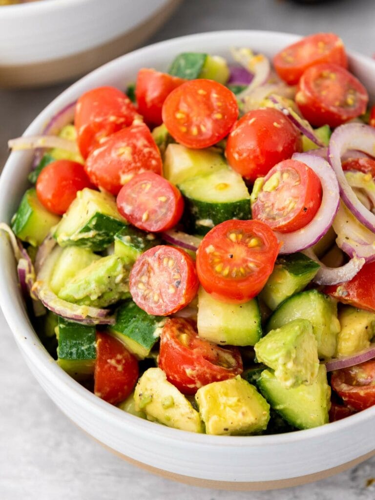 A bowl of salad containing sliced cherry tomatoes, cucumber, avocado, and red onion with a light dressing.