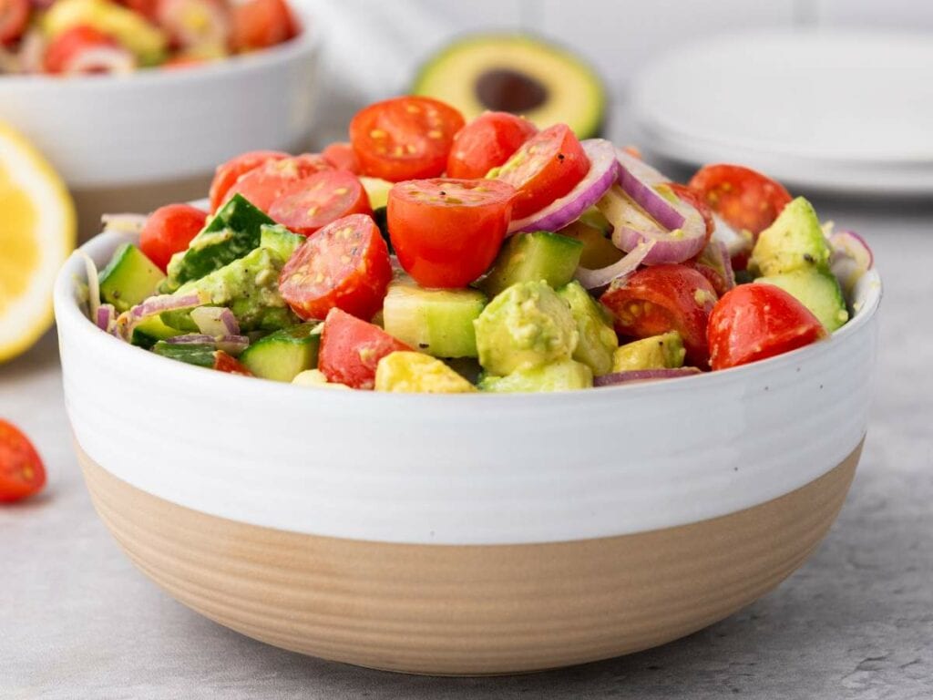 A bowl of salad containing cherry tomatoes, avocado, cucumber, and red onion slices, with more salad and avocado in the background.