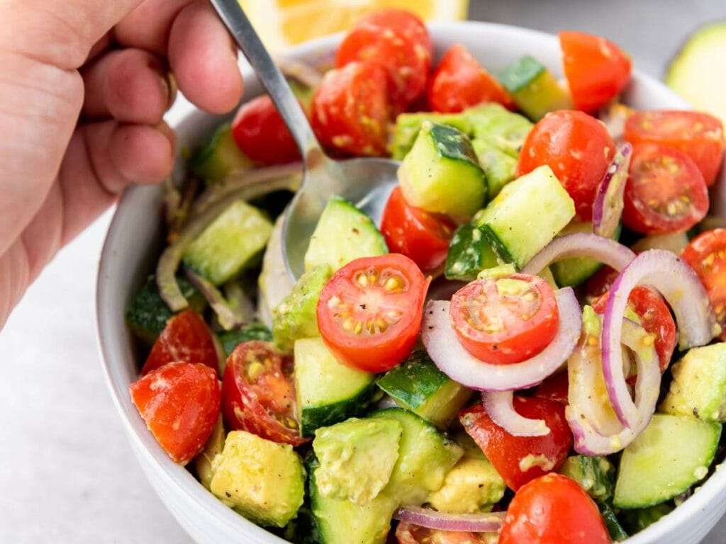 A hand holding a spoon over a bowl of salad containing cherry tomatoes, cucumber, red onion, and avocado pieces.