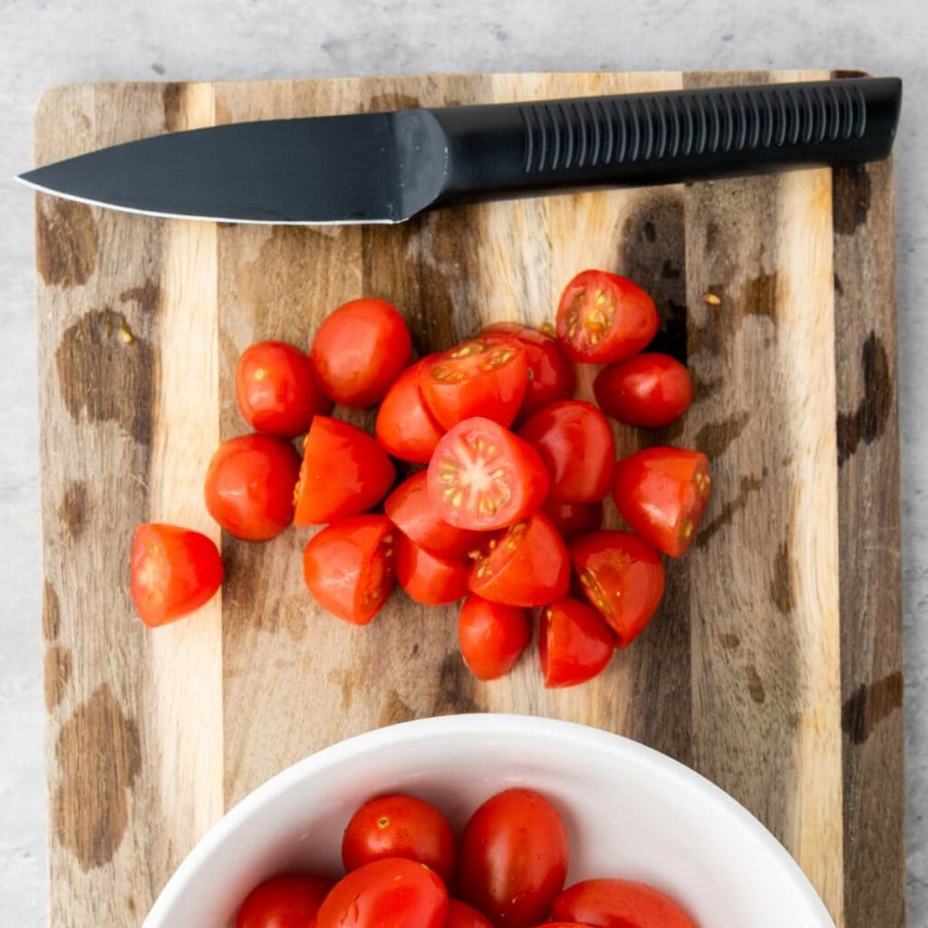 A black-handled knife and halved cherry tomatoes on a wooden cutting board, with a white bowl of whole cherry tomatoes nearby.