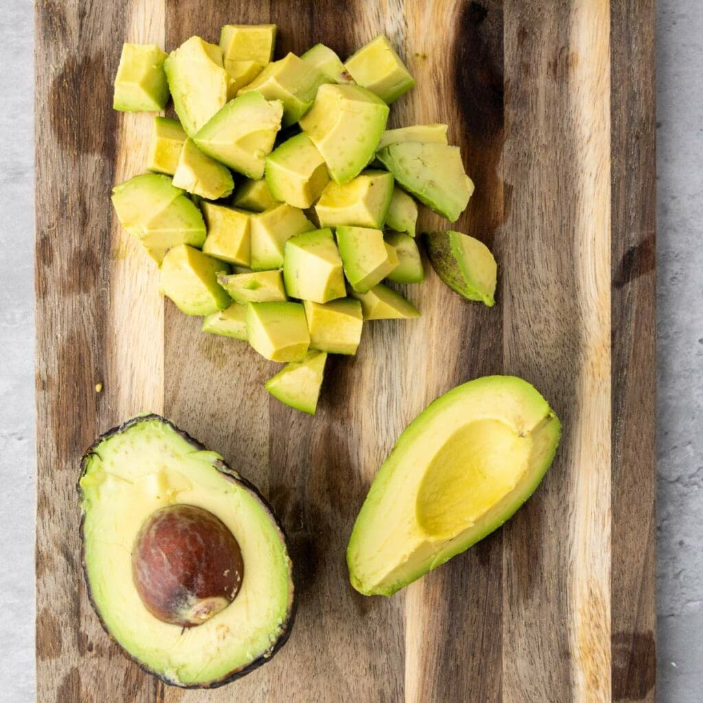 A wooden cutting board with a halved avocado, one half with the pit, one without, and a pile of diced avocado pieces.