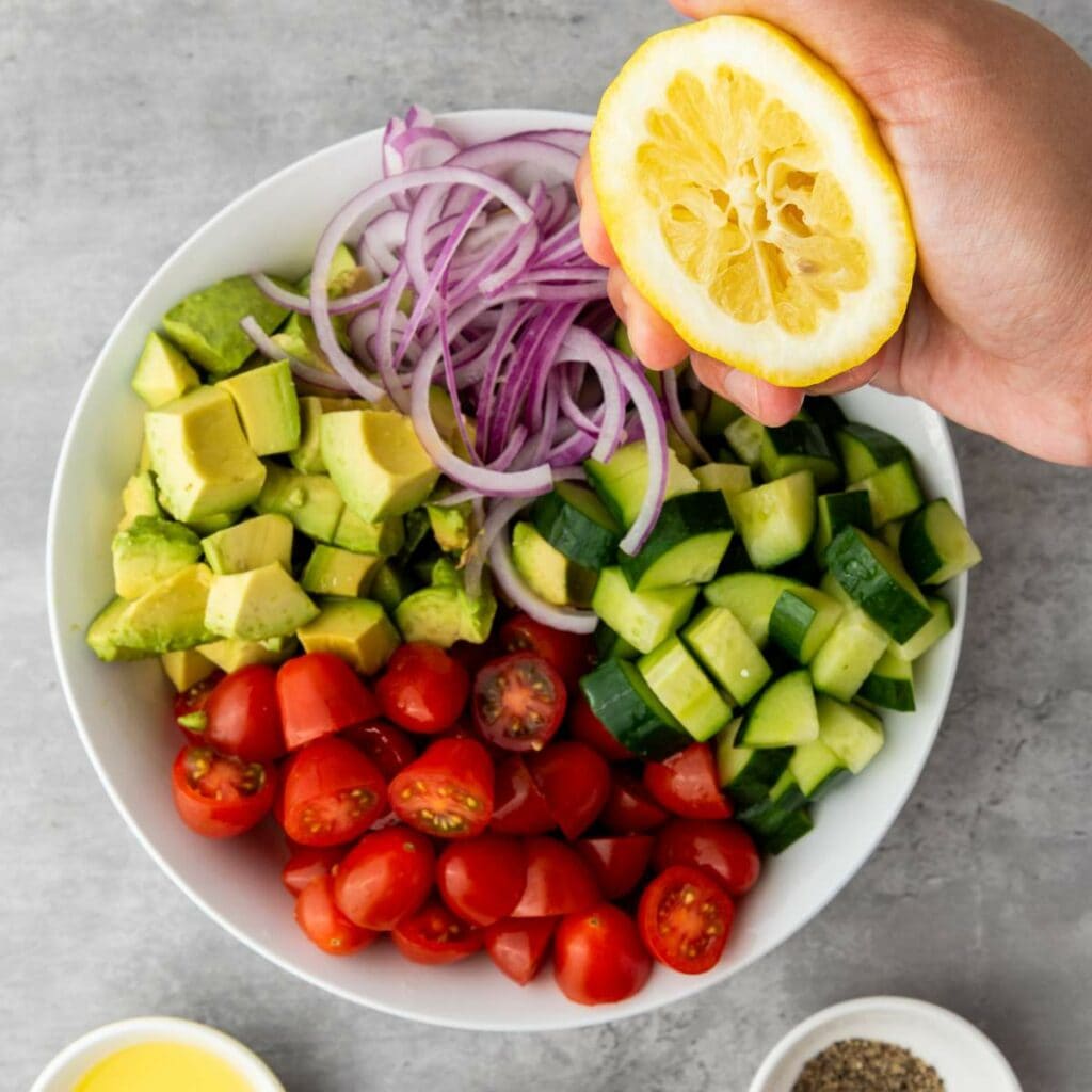 A hand squeezing a lemon over a bowl filled with chopped avocado, cucumber, cherry tomatoes, and sliced red onion.