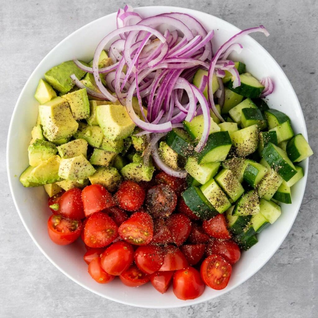 A bowl containing chopped avocado, sliced red onion, diced cucumber, and halved cherry tomatoes, seasoned with ground black pepper.