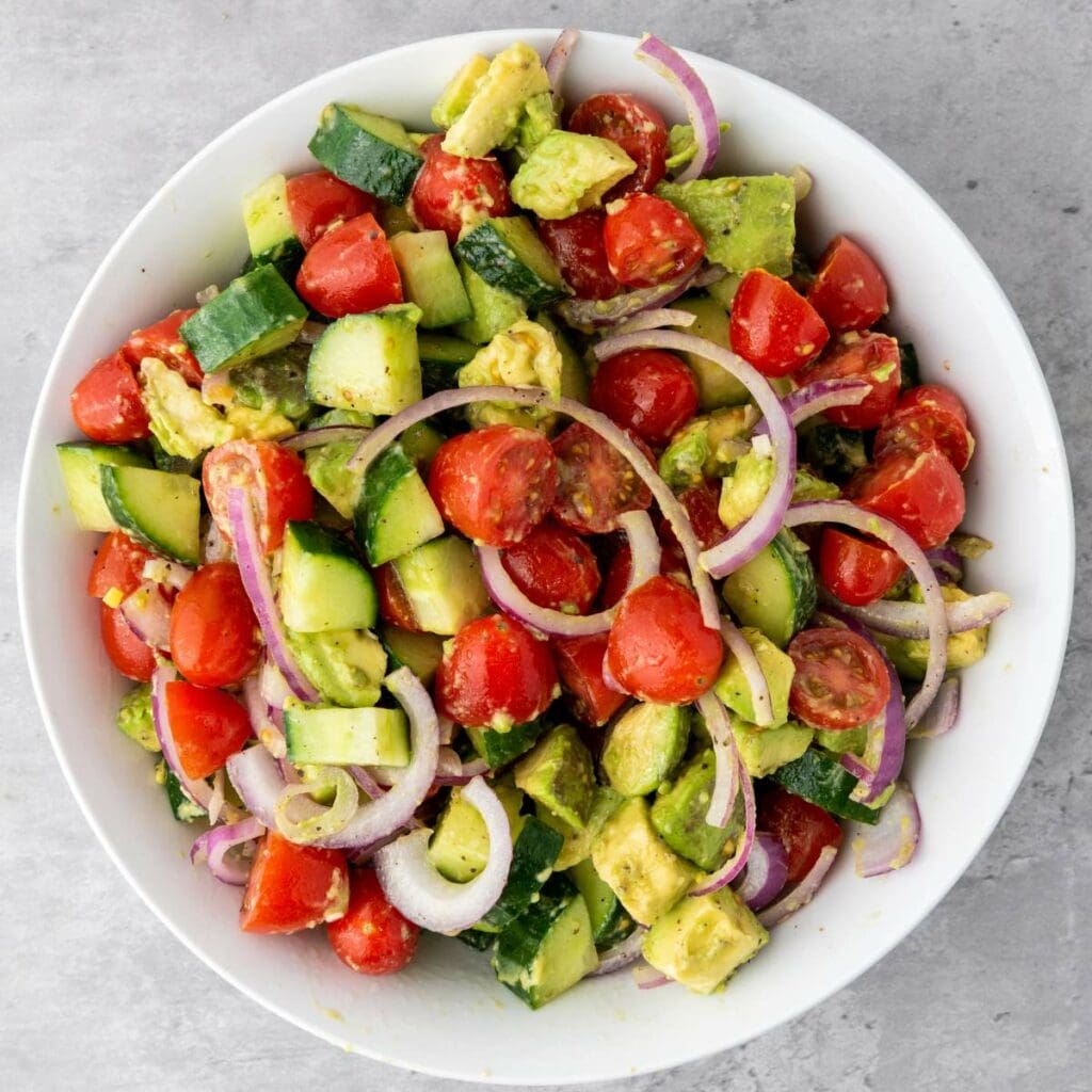 A bowl of salad with cherry tomatoes, cucumber, avocado, and red onion slices on a gray surface.
