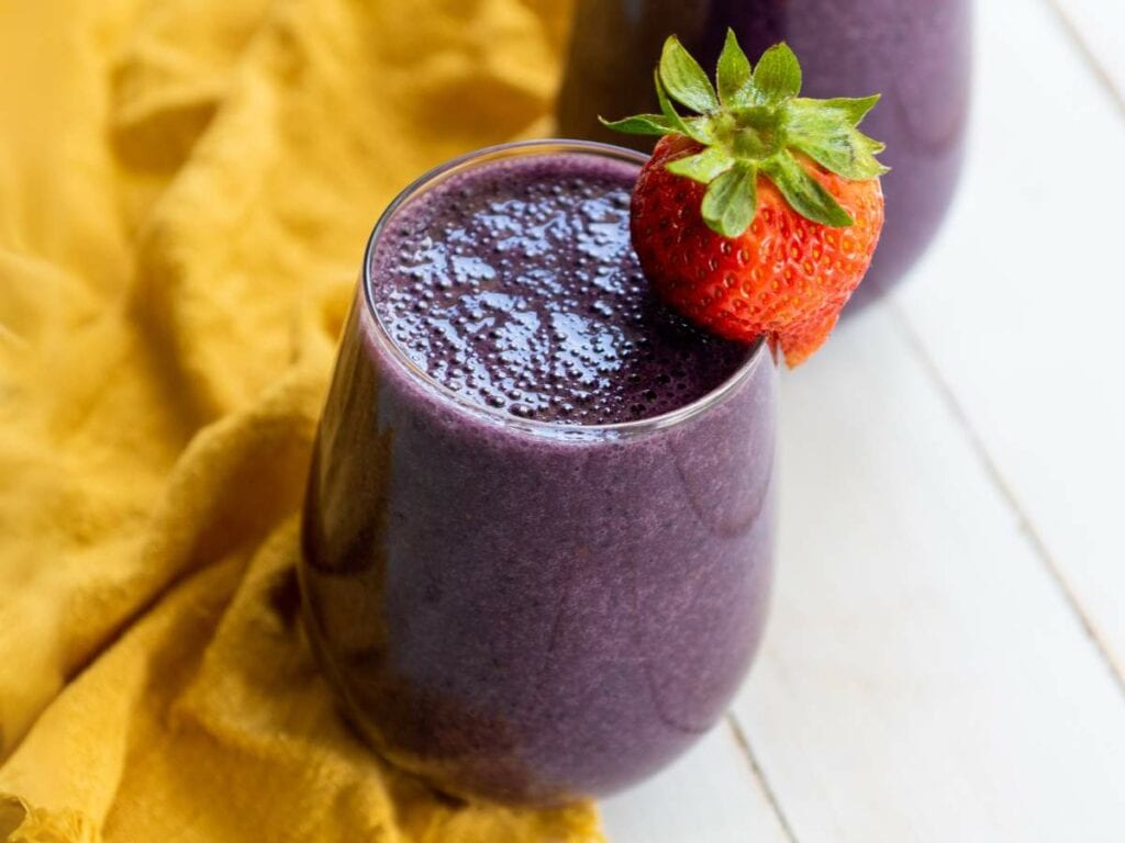 A glass of purple smoothie topped with a whole strawberry, placed on a white surface next to a yellow cloth.