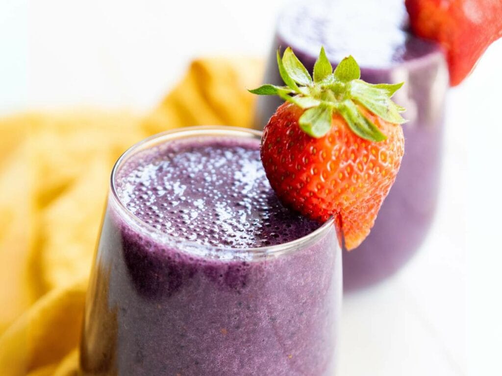 A glass of purple smoothie garnished with a fresh strawberry, with another glass and a yellow cloth in the background.