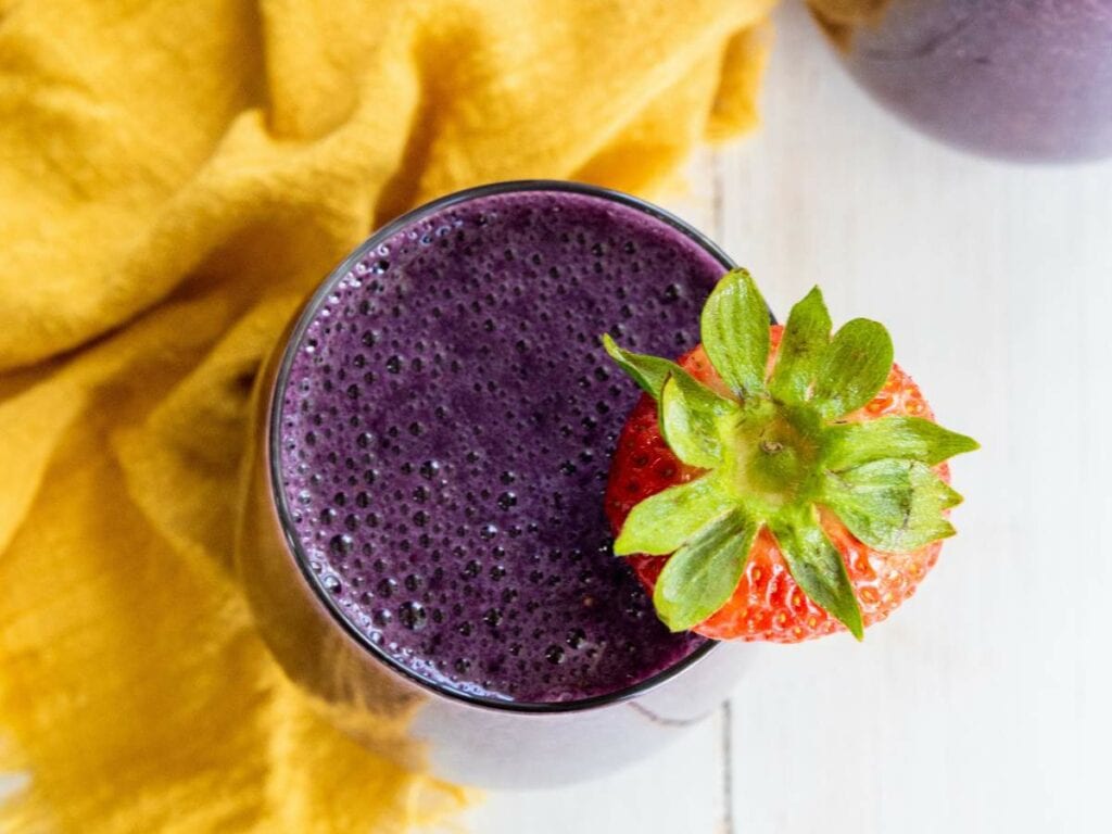 A glass of purple smoothie topped with a whole strawberry, placed on a white surface next to a yellow cloth.