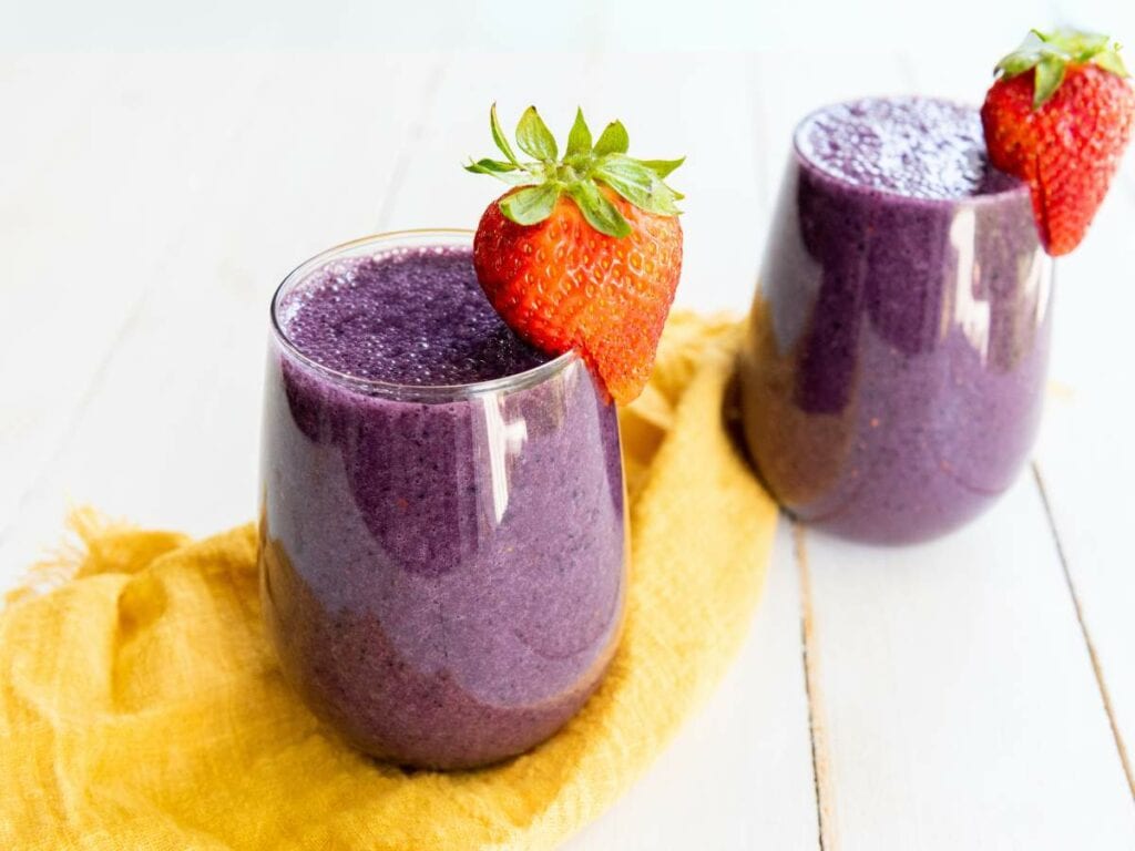 Two glasses of purple smoothie garnished with whole strawberries, set on a white surface with a yellow cloth underneath one glass.