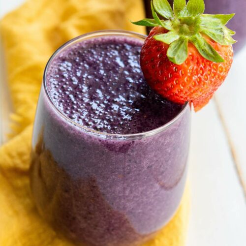 A glass of purple smoothie with a whole strawberry on the rim, placed on a yellow cloth.