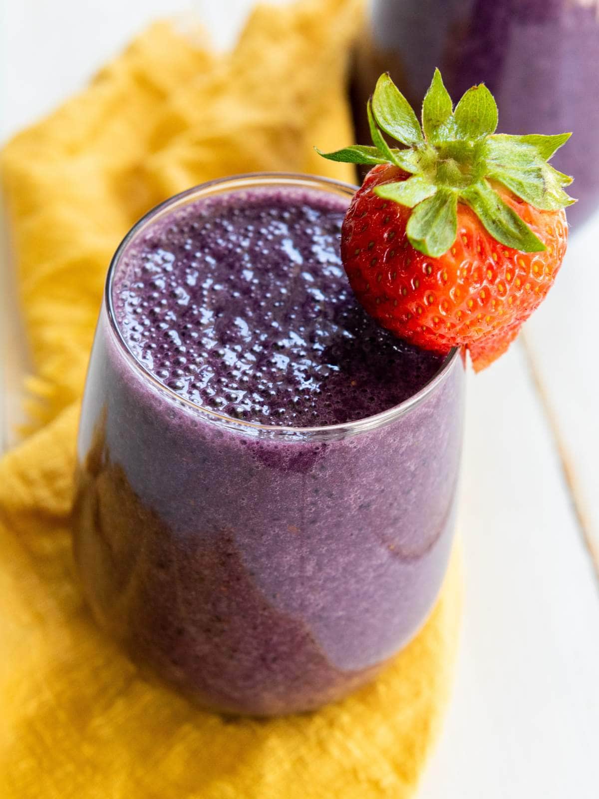 A glass of purple smoothie with a whole strawberry on the rim, placed on a yellow cloth.
